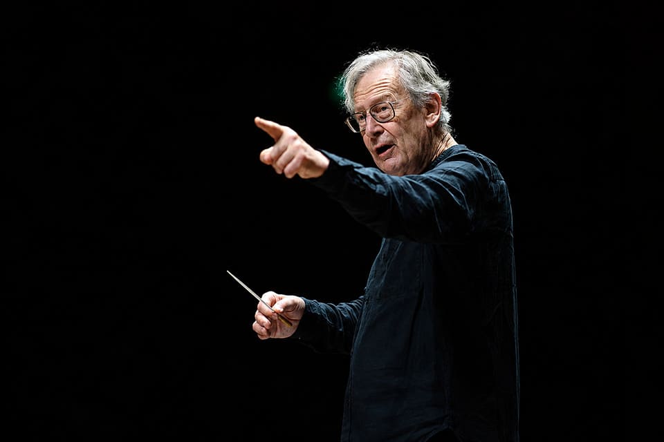 Director de orquesta señalando con una mano mientras sostiene la batuta en la otra.