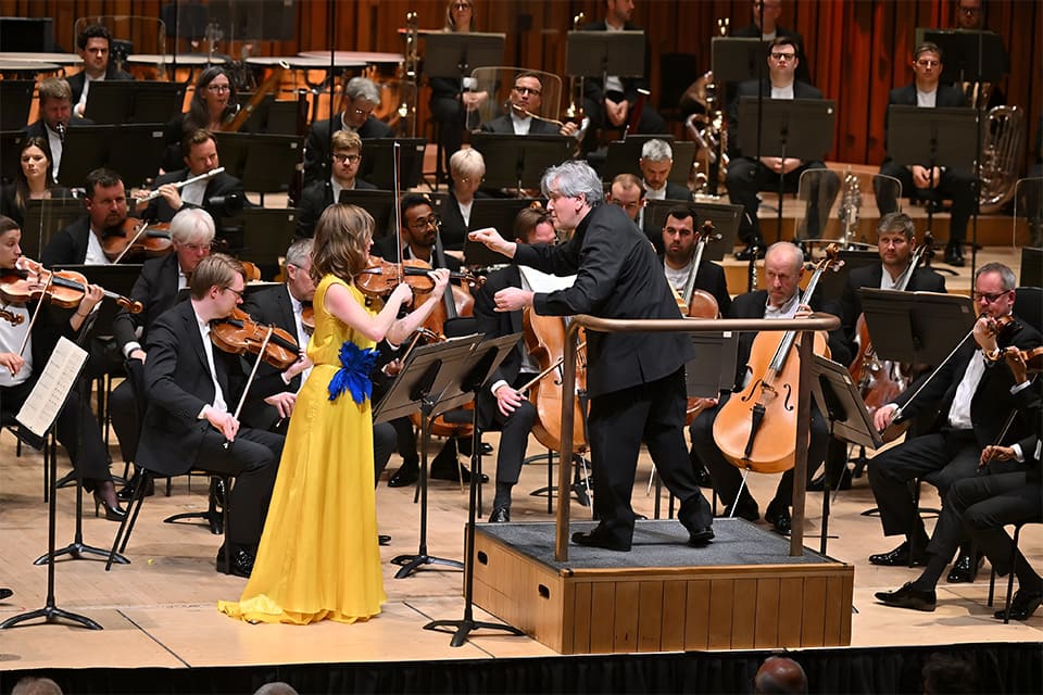 Orquesta sinfónica con solista y director, mientras varios músicos esperan su entrada al fondo.